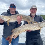 Two men holding large fish while standing on a boat in a lake with a forested shoreline in the background.