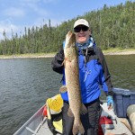 Person in outdoor gear holding a large fish on a boat, with a forested shoreline in the background.
