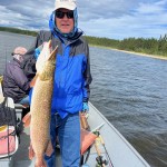 Person in a blue jacket holding a large fish on a boat with another person sitting behind, on a lake under cloudy skies.