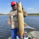 Person standing on a boat holding a large fish, wearing a camo hoodie, blue pants, and a red cap, with a lake in the background.