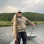 Person standing on a boat holding a large fish, with a wooded shoreline in the background.