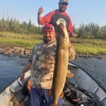 Two men on a boat; one holds a large fish while the other gives a thumbs-up. Trees and grass are visible in the background.