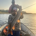 Two men in a boat are holding a large fish, possibly a pike, by a lake under a clear sky with sunlight.
