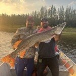 Two men in a boat holding a large fish with a forest and sunset in the background.