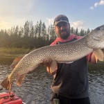 Man holding a large fish on a boat in a river, surrounded by trees and a clear sky.