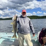 Man on a boat holding a large fish with one hand. He is wearing sunglasses, a pink cap, gray hoodie, and khaki pants.