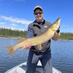 Person stands on a boat holding a large fish, with a lake and forest in the background.