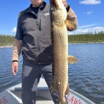 Man in sunglasses holding a large fish on a boat with a lake and forest in the background.