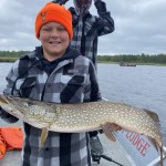 Child holding a large fish on a boat, wearing an orange beanie and plaid jacket. Another person adjusts their cap in the background.