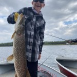 Person in a plaid shirt and sunglasses holding a large fish on a boat with a lake and cloudy sky in the background.