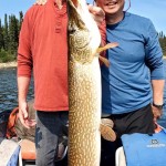 Two men in a boat holding a large fish. One is wearing a red shirt, and the other is in a blue shirt and sunglasses.