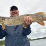 Person holding a large northern pike fish, standing outdoors with a lake and trees in the background.