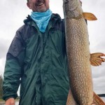 A person in a green jacket holding a large fish upright by the gills, standing outdoors with a cloudy sky in the background.