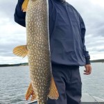 Person on a boat holding a large fish with mottled scales, against a cloudy sky and lake background.