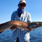 A person holding a large fish in front of a lake with a forested shoreline in the background.