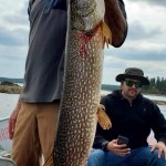 Person on a boat holding a large fish, with another person sitting nearby using a phone. Cloudy sky in the background.
