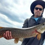 Person wearing a hat and sunglasses holding a large northern pike on a boat with a cloudy sky in the background.