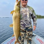 Man in camouflage attire holding a large fish on a boat with water and trees in the background.