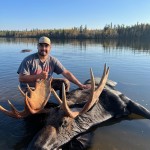 Man in a lake posing with a harvested moose. He's wearing a cap and t-shirt with trees in the background under a clear sky.
