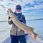 Person wearing a cap and sunglasses holds a large fish on a boat, with a cloudy sky and water in the background.