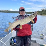 Person in a red jacket holding a large fish on a boat with a lake and forest in the background.