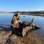 Person in camouflage kneels next to a harvested moose by a lakeshore. The landscape features trees and a clear blue sky.