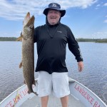 Man standing on a boat holding a large fish, with water and a distant treeline in the background.