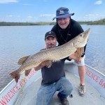 Two men on a boat proudly hold a large fish. One kneels with the fish while the other stands behind. Lake and sky in the background.