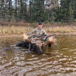 Person in camouflage with a rifle sits on a large moose in shallow water, surrounded by trees.