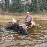 Person in camouflage sits beside a large moose in shallow water near a forested area.