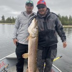 Two people in a boat holding a large fish vertically. They are wearing jackets and hats, with water and trees in the background.