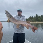 Person in a boat holding a large fish by a lake with trees in the background under a cloudy sky.