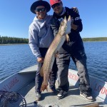 Two people on a boat hold a large fish, smiling at the camera. The background shows a lake and forest under a clear blue sky.