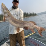 Person holding a large fish on a boat, with a lake and trees in the background, wearing a hat, sunglasses, and outdoor attire.