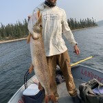 Person on a boat holding a large fish by the mouth, surrounded by fishing gear, with a lake and trees in the background.