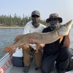 Two people on a boat holding a large fish, with trees and water in the background. Both are wearing sunglasses and face coverings.