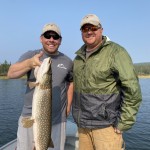 Two men on a boat, both wearing caps and sunglasses. One holds a large fish, possibly a northern pike, with a lake in the background.