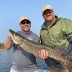 Two men wearing hats and sunglasses hold a large fish on a boat by a lake. One wears a T-shirt, the other a green jacket.