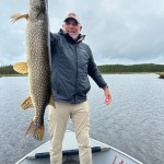 Person on a boat holding a large fish, smiling under a cloudy sky with a forested shoreline in the background.