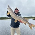 Man in a boat holding a large fish, smiling, against a cloudy sky and forested backdrop.