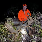 Person in an orange jacket and cap posing with a moose in a dark, wooded area.