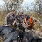 Three people in camouflage pose with a large moose on the ground in a grassy, wooded area, featuring autumn foliage.
