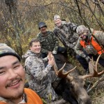 Five people in camouflage clothing smiling and posing with a moose in a wooded area.