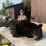 A man smiles behind a large black bear lying on a table outside, near a wooden structure and a body of water.