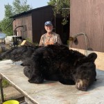 Person in camouflage stands behind a large black bear lying on a table outdoors, near a brown building and a lake.
