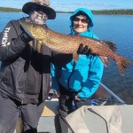 Two people on a boat holding a large fish. One wears a hat and mask, the other wears a blue jacket. Lake and trees in background.