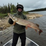 Person standing on a boat holding a large fish, with a wooded shoreline and cloudy sky in the background.