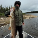 A person in a green jacket holds a large fish on a boat near a wooded shoreline under a cloudy sky.