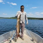 Person standing on a boat holding a large fish. A fishing net and oar are on the deck, with a lake and trees in the background.
