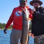 Two people on a boat holding a large fish, with a lake and trees in the background.